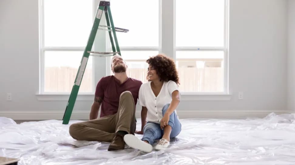 Two people sit on plastic drop cloth in a white room with a ladder.