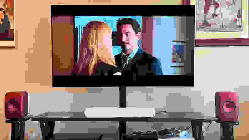 The Sonos Ray sits on a brown TV console between red speakers with Iron Man shown on screen.