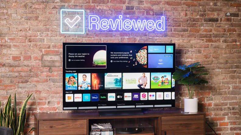 A webOS screen on an LG C2 OLED sits on a wooden table in front of a brick wall, with an outline neon sign hanging above it.