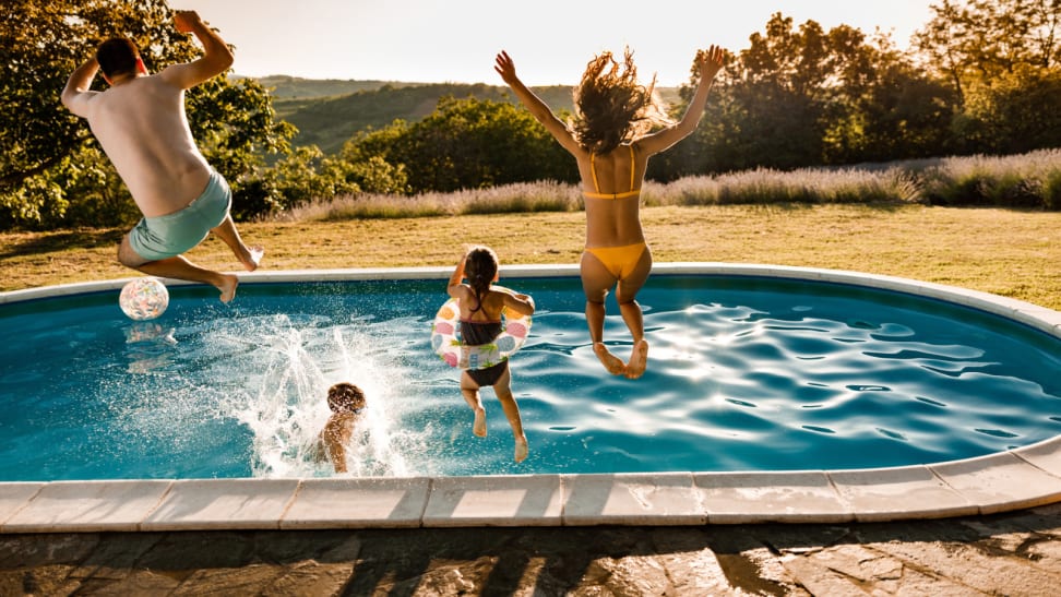A family of four jumping into an in-ground, backyard swimming pool at dusk