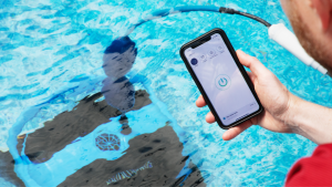 Dolphin Nautilus CC Plus Wi-Fi robot pool vacuum in a swimming pool with a person holding a smartphone showing the app in the foreground.