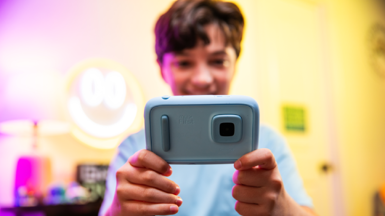 A child holding a light blueCapture every moment of fun with the kid-friendly myFirst camera in a playful, durable design. camera with a smile, captured in a colorful and cheerful room featuring a glowing smiley face sign in the background.