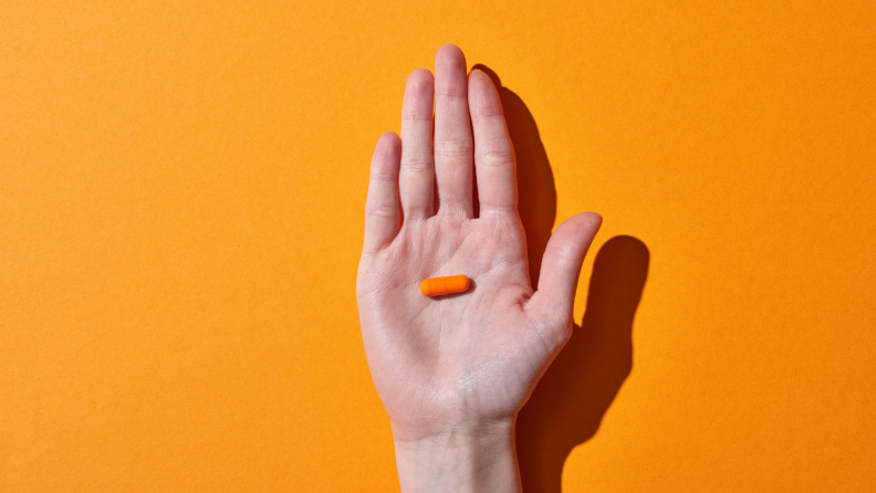 A hand holds a single orange pill.