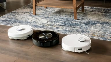 Three robot vacuum and mop hybrids lined up on a brown floor next to a rug.