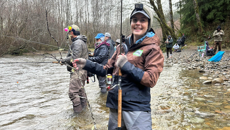 A woman wearing Simms fishing clothes and holding a fishing rod, in a river, next to other fisherman.