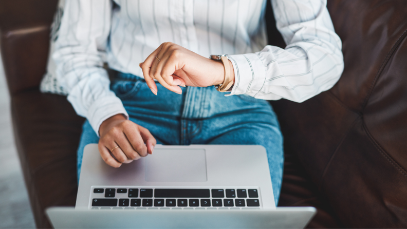 Woman sitting down and checking watch with laptop on lap.