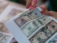 Two hands use scissors to cut U.S. dollar bills out of a printed piece of paper.