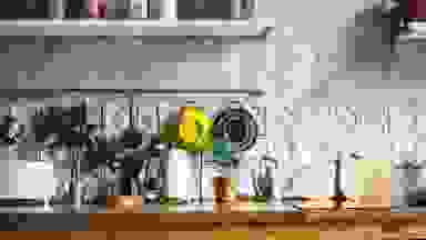 阳光普照的厨房台面摆满了植物和小电器，包括一个黑色的moka锅、木制砧板、一个白色的双片烤面包机、一个白色的电水壶，以及挂在上面的两个滤锅，它们挂在白色的纹理墙上。