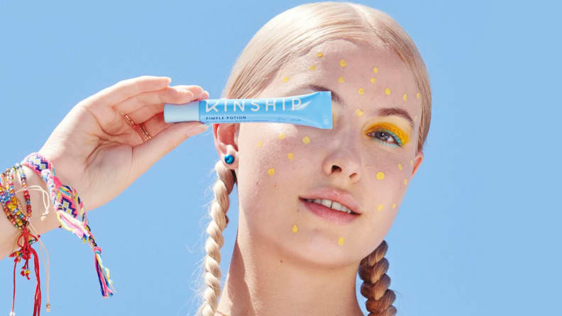 A person with long blond hair in braids on either side of their head holds up a blue squeeze tube in front of their eye while having yellow dots all around their face.
