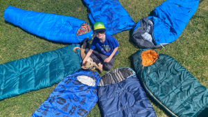 A round of kids' sleeping bags, including Kelty Mistral, REI Kindercone, L.L. Bean Adventure Kids, and an ANJ on the grass with a child and puppy