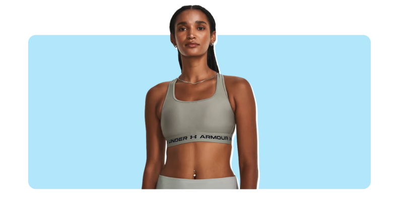 Model wearing gray Under Armour Mid Crossback Sports Bra with black words printed on the elastic band.