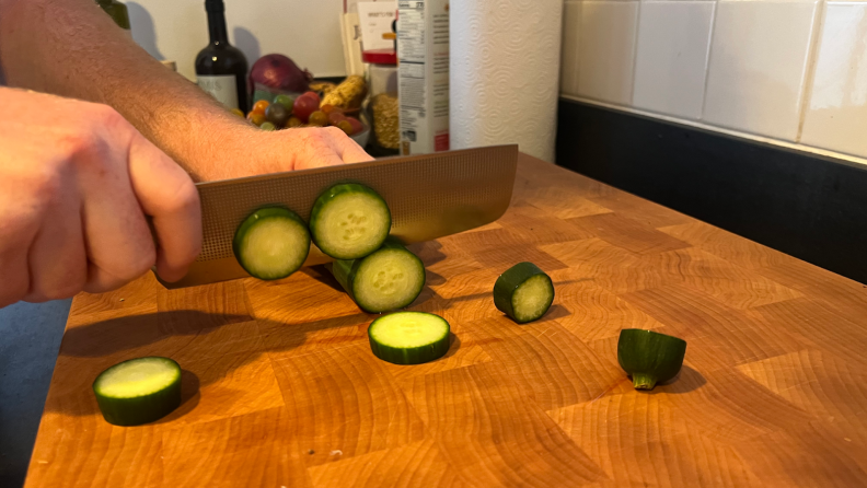 A person using the Milk Street Nakiri knife slicing a cucumber into smaller slices on top of a wooden cutting board.