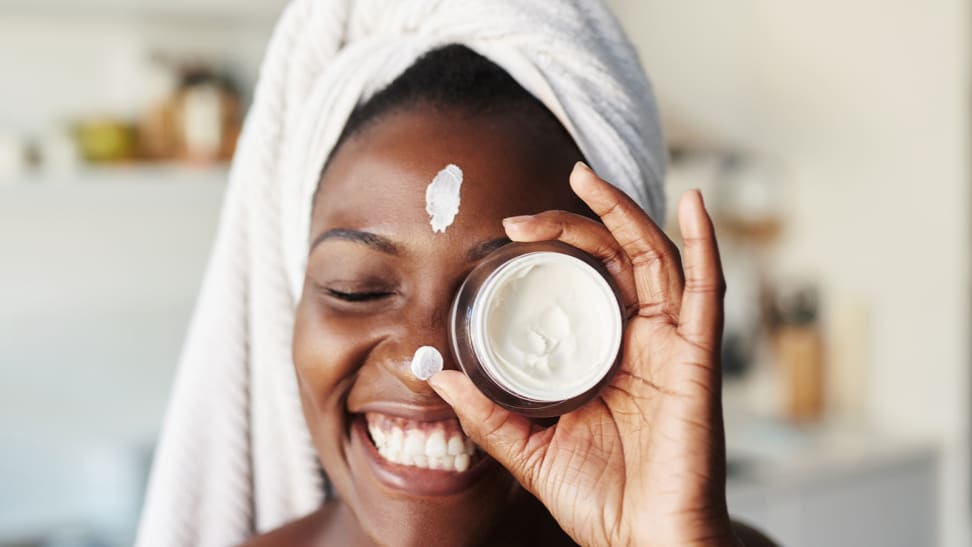 A woman wearing a towel on her head and smiling as she holds up a jar of cream in front of her eye. She has a strip of moisturizer on her forehead and on her nose.