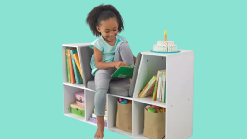 A child smiles as she sits and reads in a small reading nook that has book shelves and seating.