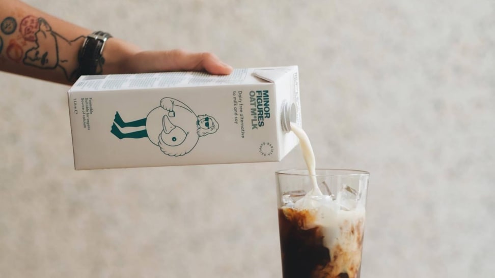 一个纹身的人将燕麦牛奶从纸箱中倒入一杯冰咖啡中。