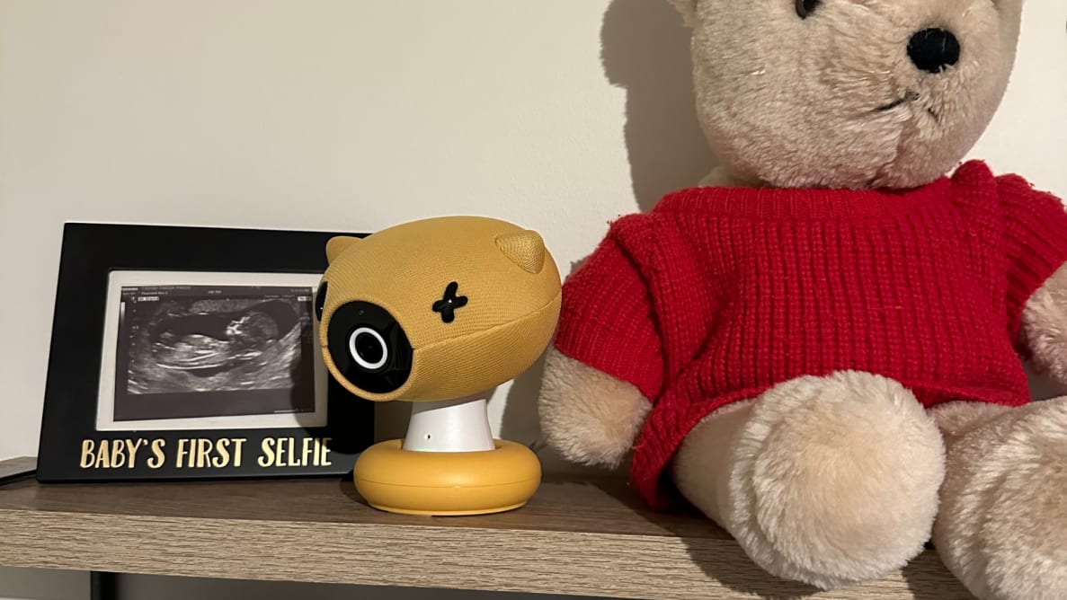 A yellow baby monitor sits on a shelf next to a brown teddy bear wearing a red sweater