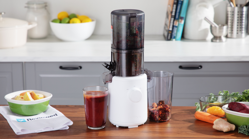 The Hurom H310A sits in the middle of a wood counter with a cup full of red juice on one side and juicer waste on the other. Produce sits on the counter.