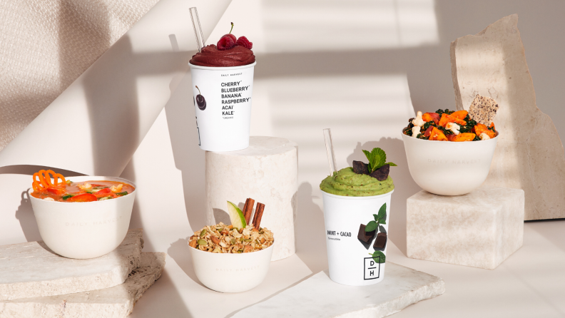Two Daily Harvest smoothies and three bowls on white pedestals and a white background.