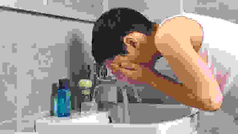 A teen boy washes his face over the sink.