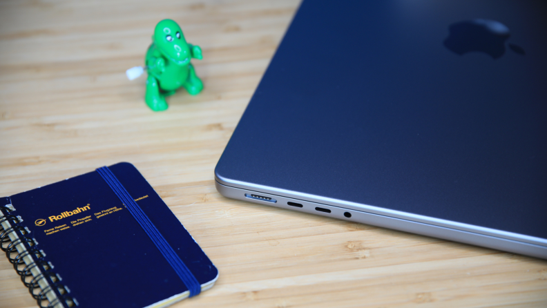 The Apple MacBook Pro 14 (2023) is closed sitting on a wood table, next to a green, toy dinosaur, and a notebook.