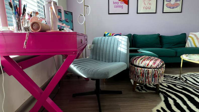 Blue Pukami Armless Desk Chair sitting in front of pink desk next to a colorful ottoman inside of a colorful living room on a sunny day.