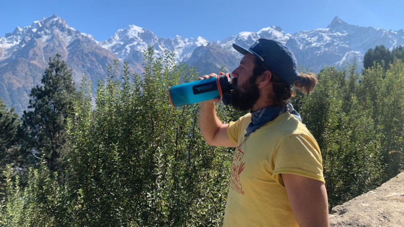 A bearded man in a yellow shirt, bandana, and hat drinks from a Grayl Geopress filtered water bottle.