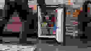 A man steps into frame to access the interior of an open Insignia NS-CF26BK9 mini fridge. It's stocked full of food and drinks.