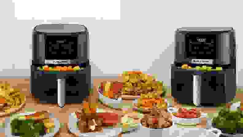 Black and stainless steel Kalorik 5-Quart Digital Air Fryer with basket filled with potato tator tots and surrounded by assorted cooked foods inside of white dishes.