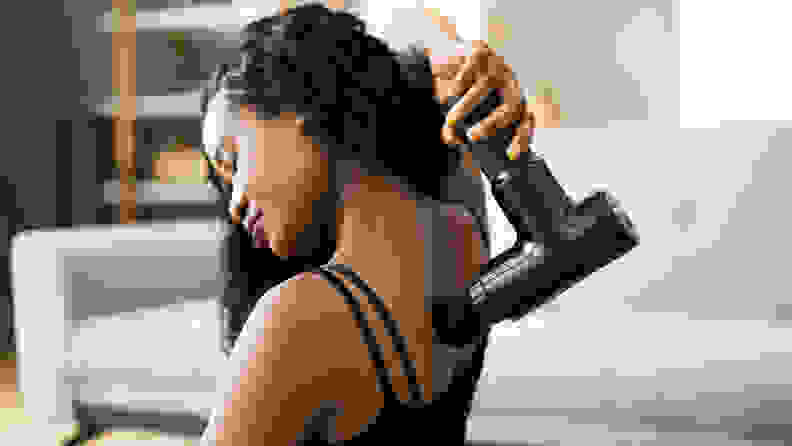 A woman using a massage gun on her upper back muscles.