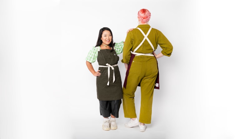 Two people wearing cross-back aprons and posing in front of a white background.