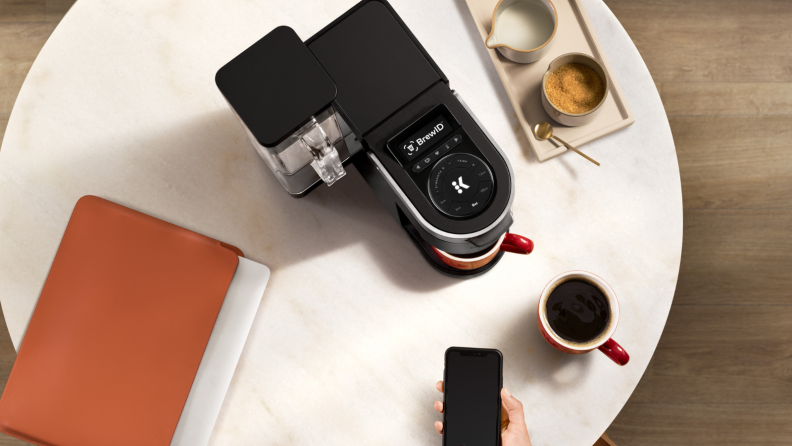 On a white marble coffee table, there's a Keurig K-Supreme Plus Smart pod coffee maker. Next to the machine, there's a coffee mug, a note book, and a tray of drinks. A person is pointing a smartphone at the machine, hinting the coffee maker's smart technology.