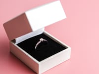 This is an engagement ring in a white box against a pink background.