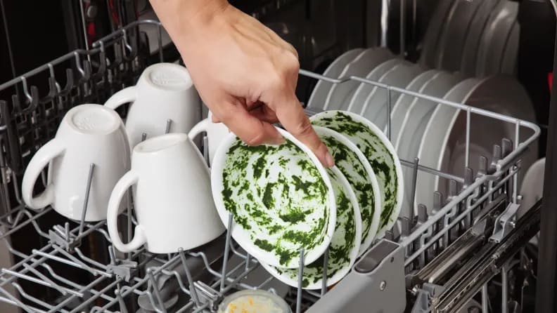 A person loading a bowl covered with spinach puree into a dishwasher that also has plates and mugs in it.