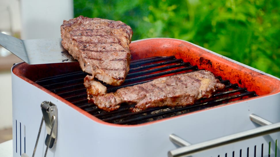 A person is flipping a piece of steak on a charcoal grill using a spatula. There's another piece of steak on the grill.