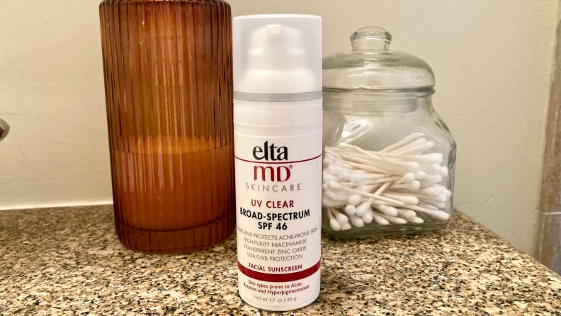 A white cylindrical tube of sunscreen sits on a granite sink with soap and a container of cotton swabs behind it.