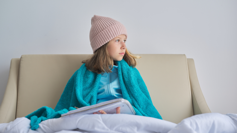 Child in bed looking to their left with beanie, coat, and blanket