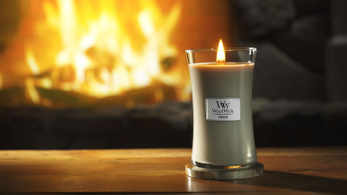 A lit WoodWick candle sits on a table in front of a fireplace.