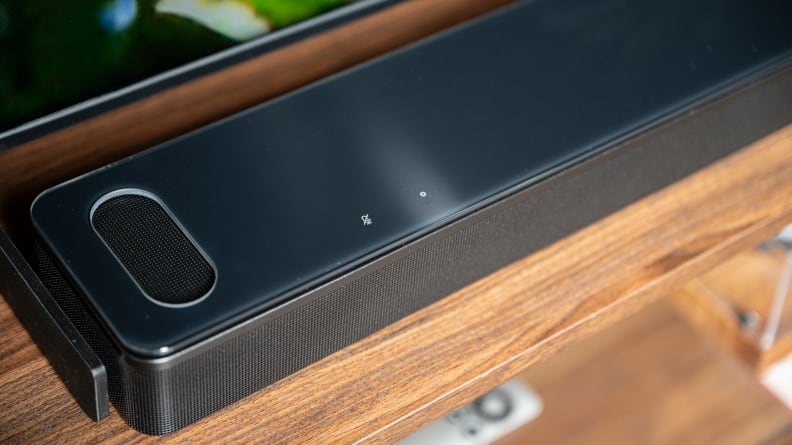 The glass-topped, all-black Bose 900 soundbar sits atop a wooden TV console with cuts in the top revealing an upward firing driver.