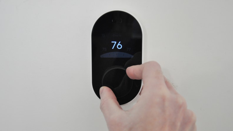 Black Wyze Thermostat mounted against white wall. Display shows "76," and a hand is adjusting the dial on the thermostat's face.
