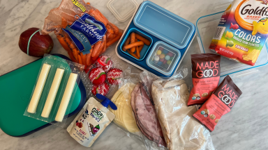 Bento boxes, cheese sticks, carrots, an apple, an assortment of deli meats and packaged snacks laid out on a marble counter.