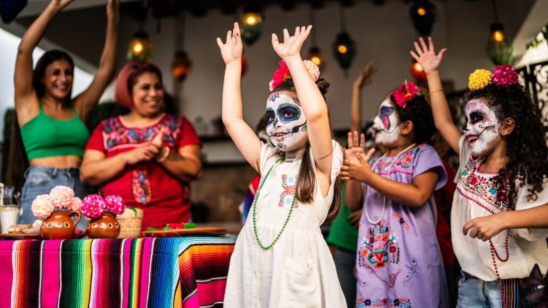 Small children with painted faces smile with their hands in the air at a Dia de Los Muertos event outdoors.