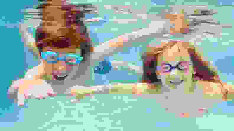 Two kids swim under water smiling in their swimming goggles