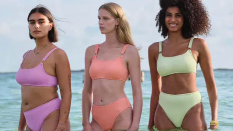 Three models wearing purple, orange and lime green two-piece swimsuits in ocean.