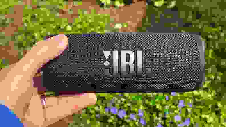 A black, cylindrical Bluetooth speaker is held in a hand above greenery with a JBL logo in the front.