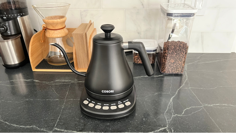 Cosori Electric Gooseneck Kettle sits on a black counter with a Chemex and containers of coffee beans in the background.