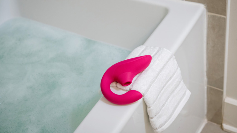 A Womanizer Blend vibrator in pink resting on a bath towel on the rim of a bathtub.