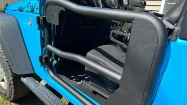Angled outside view of a Rugged Ridge Fortis Tube Door installed on the driver's side of a light blue Jeep.