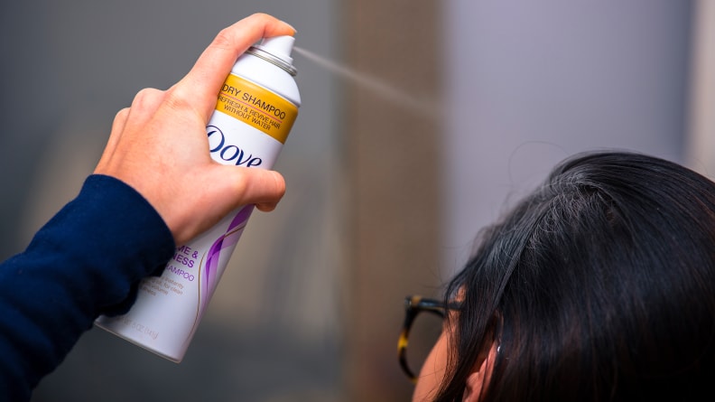 A person with dark, hair sprays the Dove Volume & Fullness Dry Shampoo into their hair from several inches away.