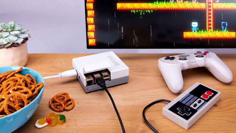A tiny gaming console with a controller attached on a desk with a monitor and a bowl of pretzels