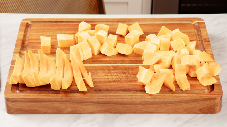 Butternut squash sitting on top of wooden cutting board after being cut into various shapes before being cooked.
